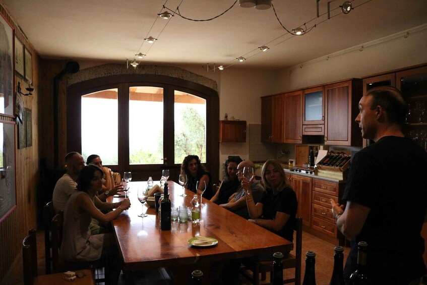 A group of people seated around a wooden table in a kitchen with wooden cabinets and a large window. 2