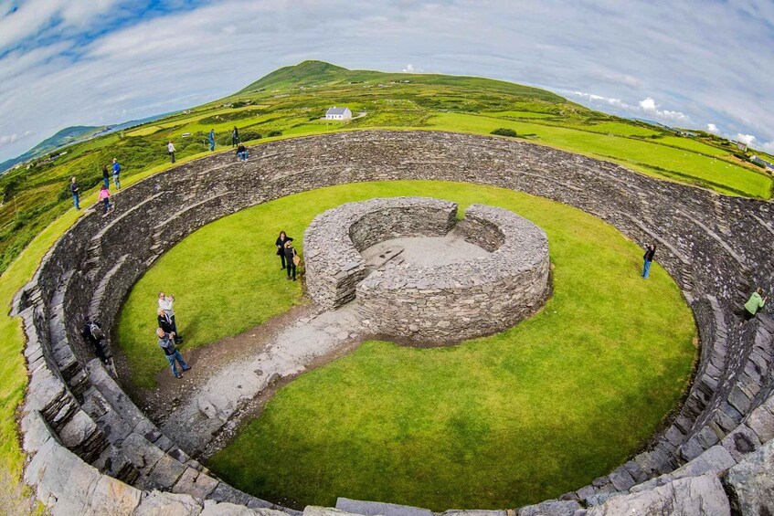 A stone circular structure with a grassy interior, surrounded by a stone wall, set in a hilly landscape with a small white building in the distance. 1