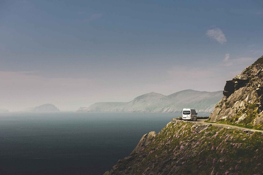 A white van on a narrow mountain road with a view of the ocean and distant mountains. 5