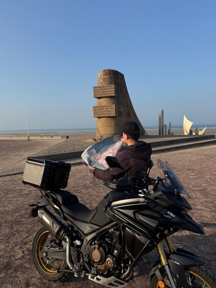 A person sitting on a motorcycle with a map, near a monument and beach. 3