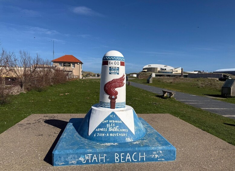 A monument with a winged column, 'KM 00 VOIE DE LA LIBERTÉ 1944' and 'UTAH BEACH' inscribed, with a building and a road in the background. 2