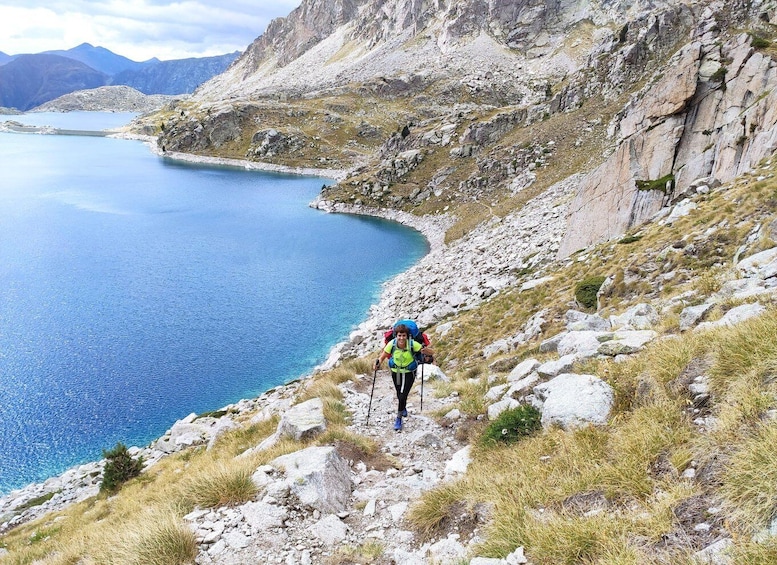 A hiker on a rocky path beside a calm lake with mountains in the background. 2