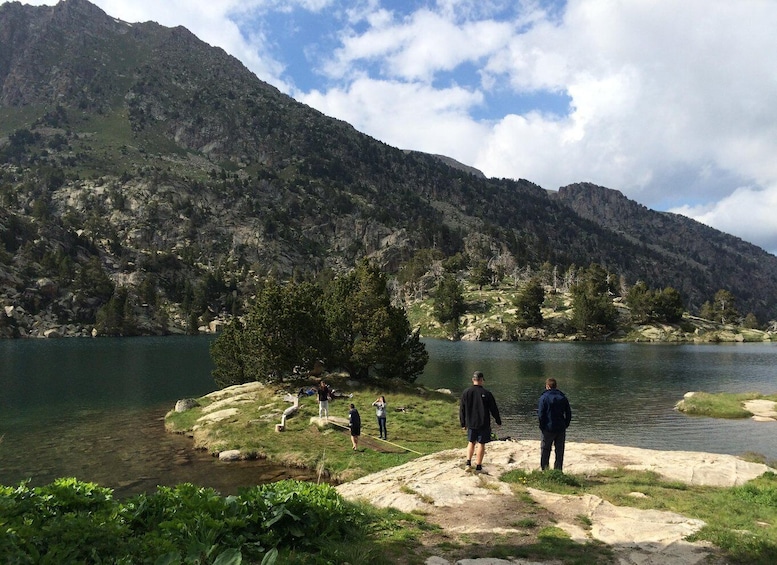 A group of people standing on a rocky shore by a lake, with mountains in the background. 1