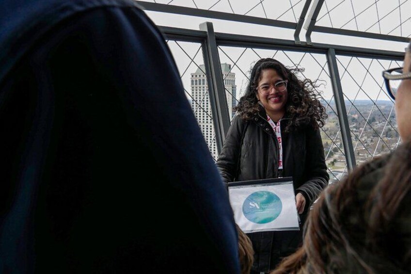 A person holding a sign with a circular logo, standing in front of a large window with a cityscape view. 4