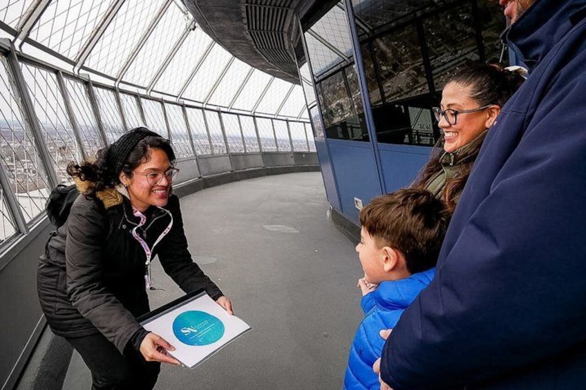 A group of people inside a glass-enclosed structure, possibly a viewing platform, with a cityscape in the background. 3