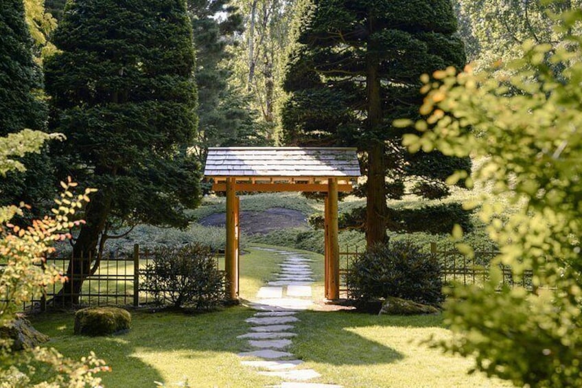 A wooden gazebo in a park with a stone pathway leading to it, surrounded by trees and shrubs. 2