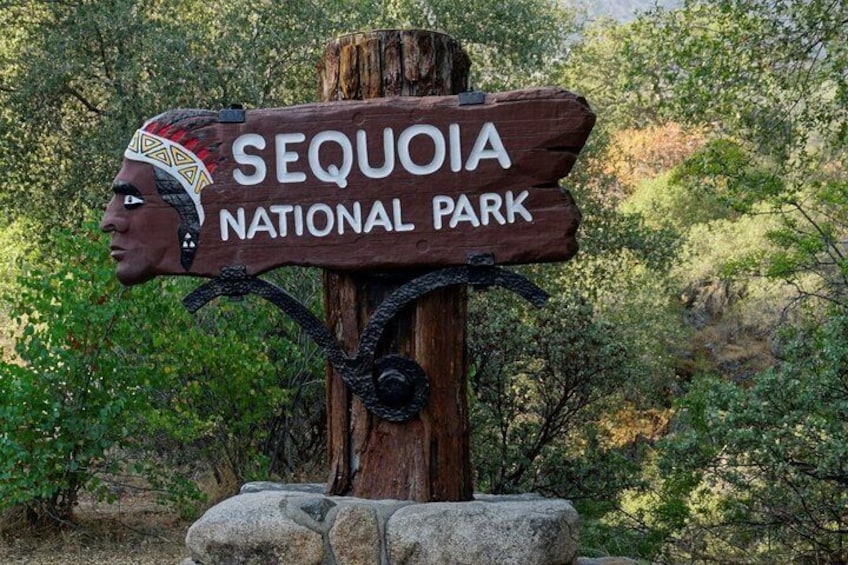 A wooden sign with 'SEQUOIA NATIONAL PARK' written on it, mounted on a post with a carved Native American head at the top. 1