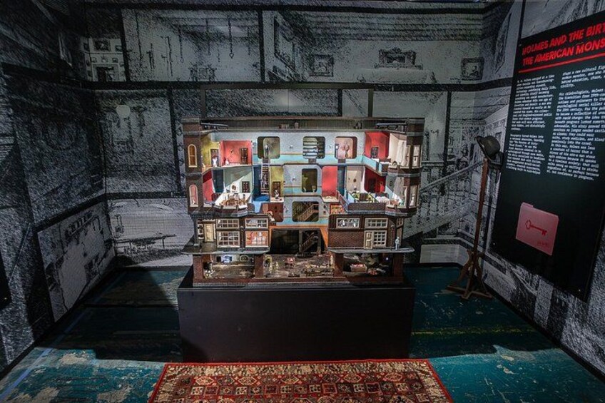 A model of a multi-story building with various rooms and furniture, displayed in a room with a patterned carpet and a wall with text. 2
