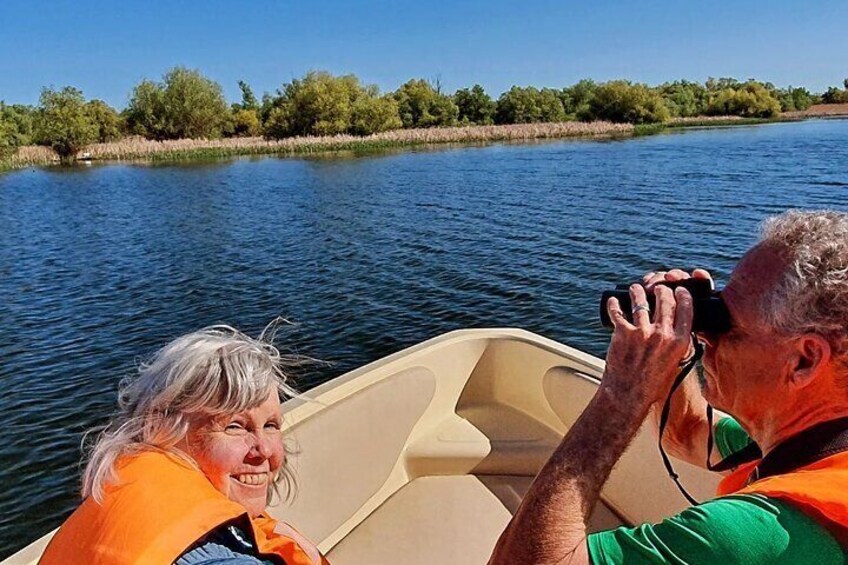 A person in a boat wearing an orange life jacket, another person in a green shirt, and a calm river with trees in the background. 1