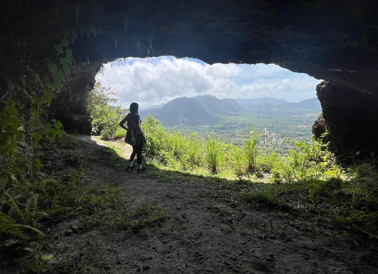 A person walking on a dirt path through a natural cave opening, with a view of mountains and a cloudy sky. 5