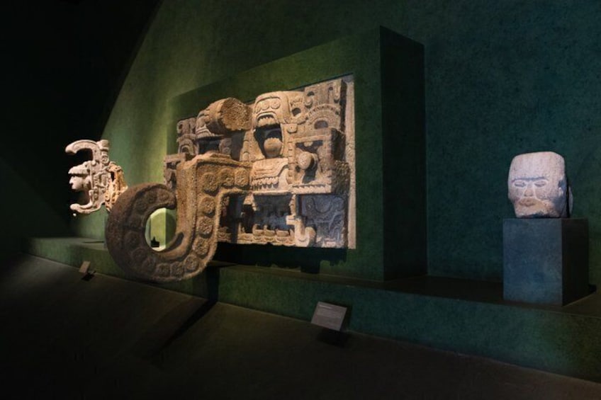 A stone carving of a figure with a large circular base, a smaller figure beside it, and a stone head on a pedestal. 3