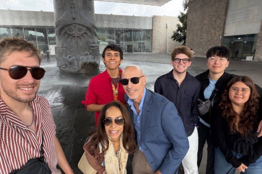 A group of people posing in front of a large stone sculpture with a face, in an outdoor setting with a building in the background. 2