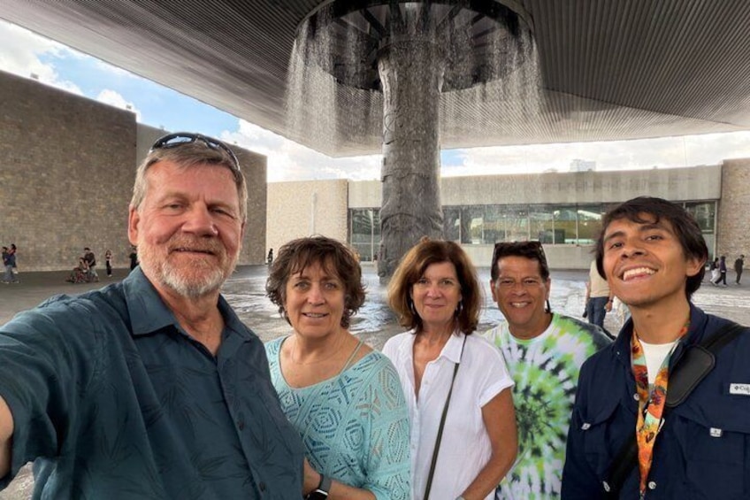 A group of people posing in front of a modern building with a large column and a water feature. 1