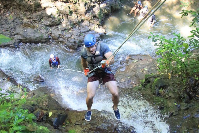 A person rappelling down a rocky cliff with a rope, surrounded by greenery and a flowing river. 2