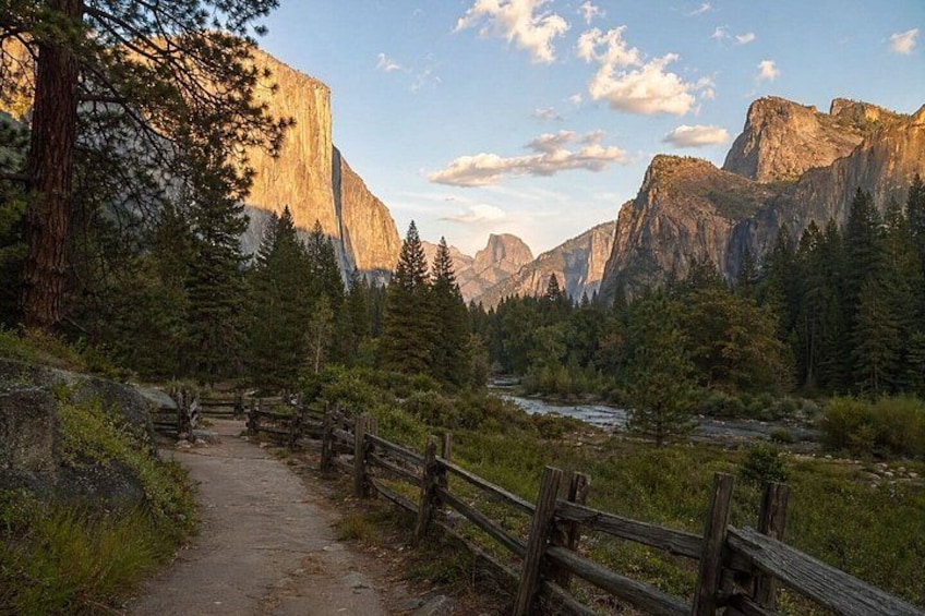 A forested mountain path with a wooden fence, a river, and towering cliffs. 4