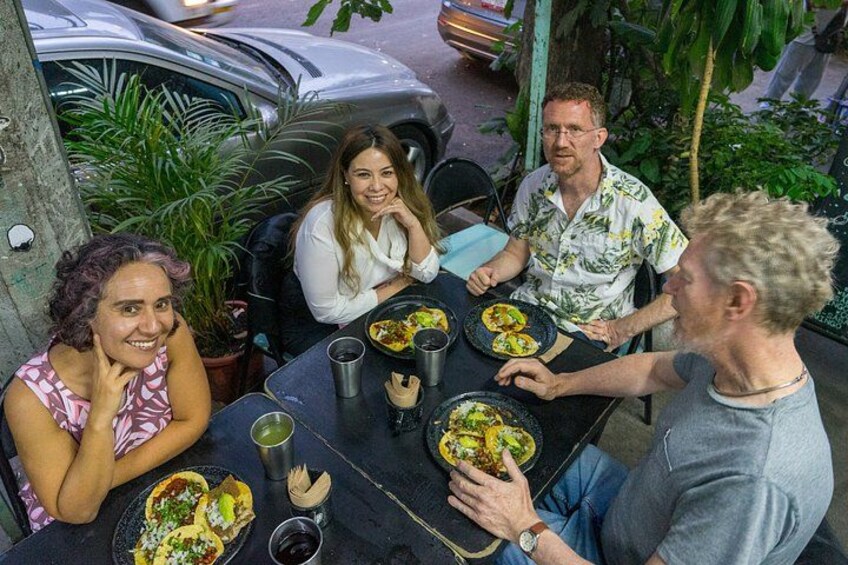 A group of people dining outdoors at a table with plates of food, cups, and a bottle. 1