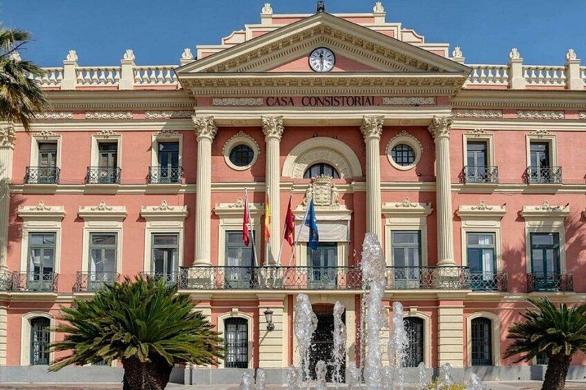 A pink building with columns, a clock, and flags. 2
