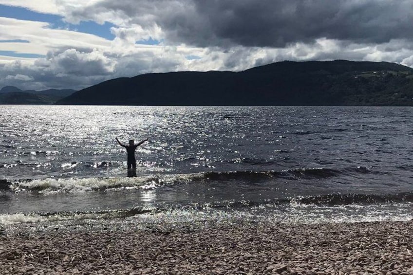 A person standing in shallow water with arms raised, a rocky shoreline, and mountains in the background. 3