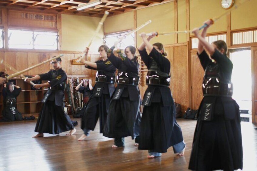 A group of people in traditional attire practicing with wooden staffs in a wooden-floored room. 1