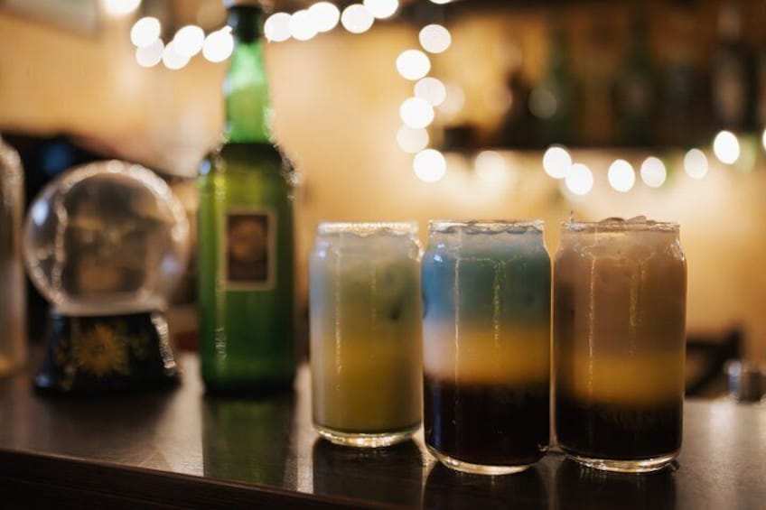 Four layered drinks on a bar counter with a blurred background of lights and a green bottle. 3