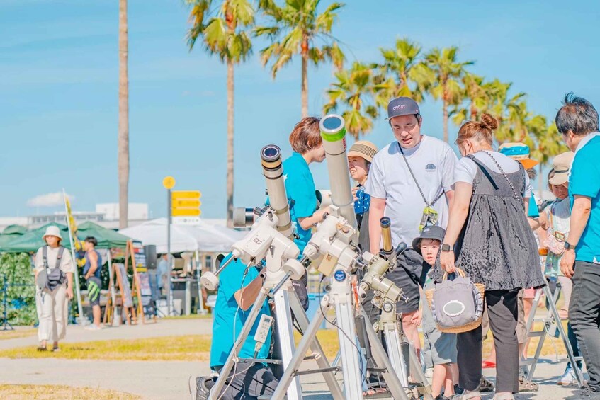 A group of people observing telescopes at an outdoor event with palm trees in the background. 2