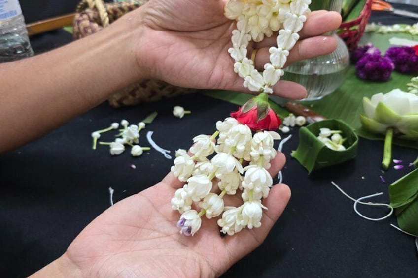 Hands holding a garland of white flowers, with a red rose and green leaves in the background. 3