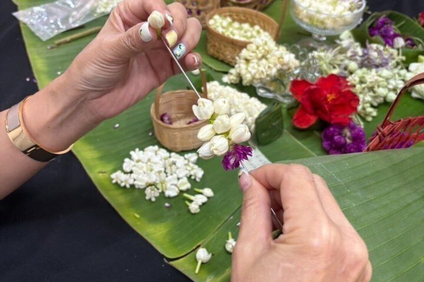 Hands threading flowers onto a string, with various flowers and a green leaf in the background. 4