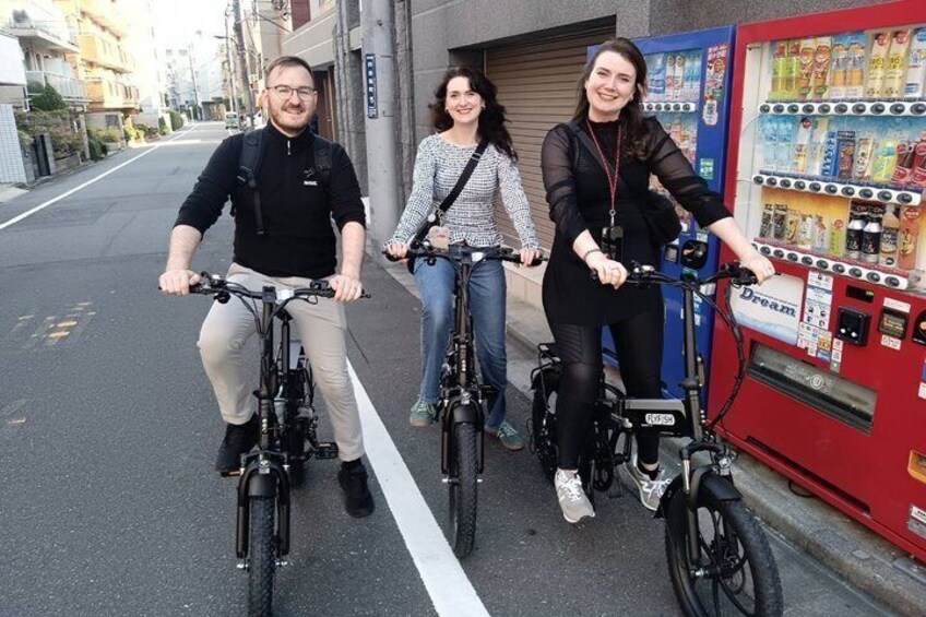 Three people on bicycles on a city street with a vending machine. 2