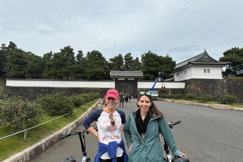 Two people on bicycles, a traditional Japanese building, and a stone wall in the background. 3