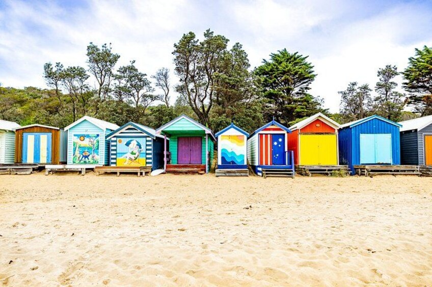 A row of colorful beach huts with unique designs, set against a sandy beach with trees in the background. 4