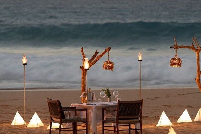 A beach dinner setup with a table, chairs, and decorative lights. 1