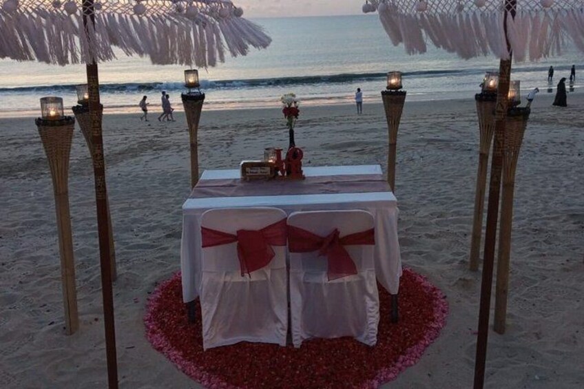 A beachside dining setup with a table adorned with a white tablecloth and red ribbons, surrounded by wicker chairs and lit lanterns. 3