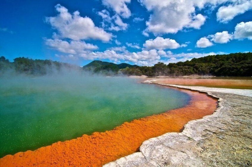 A geothermal hot spring with green and orange water, surrounded by forested hills. 1