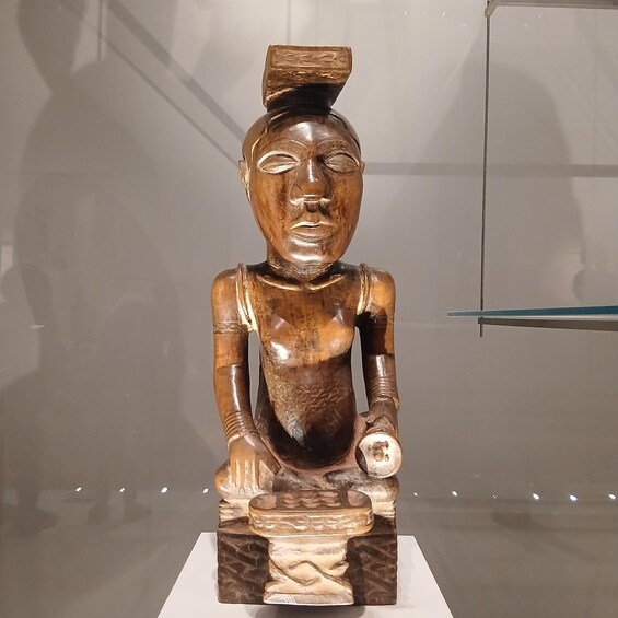 A wooden sculpture with a seated figure, a bowl on its lap, and a headdress on its head. 3