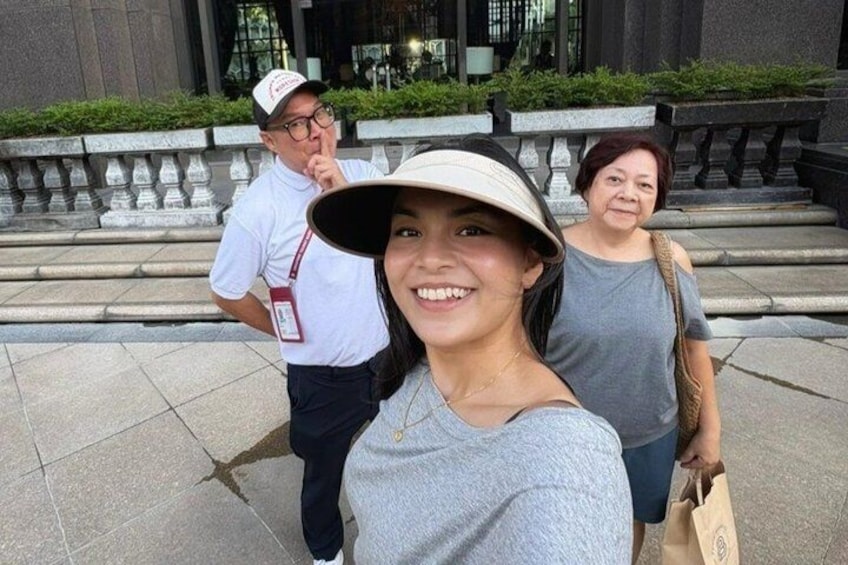 A person in a grey top and white hat, another in a grey top, and a third in a white shirt with a red badge, standing on a paved area with steps and greenery in the background. 1