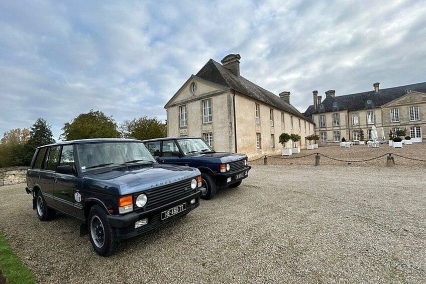 Two vintage cars parked in front of a large, historic building with a gravel driveway. 4