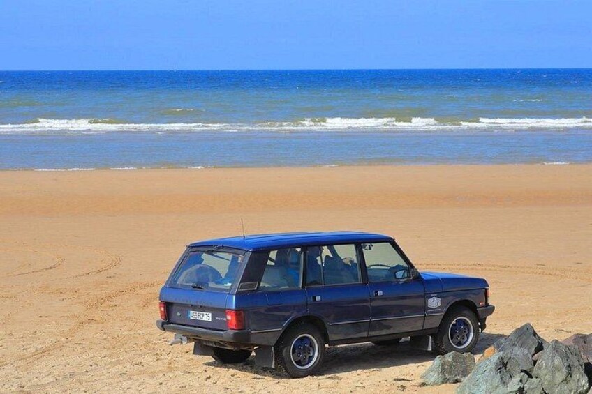 A blue car parked on a sandy beach with the ocean in the background. 1