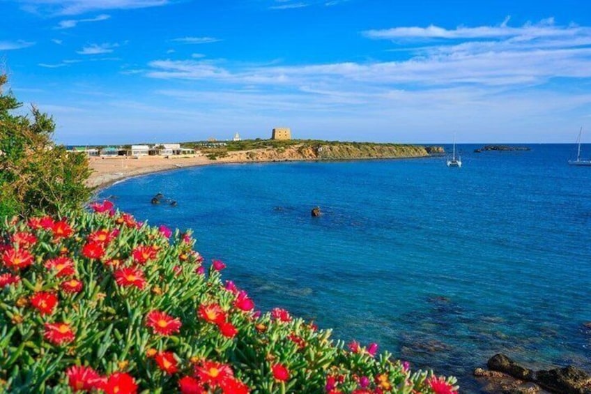 A coastal landscape with clear blue waters, a sandy beach, and a distant rocky outcrop. 5