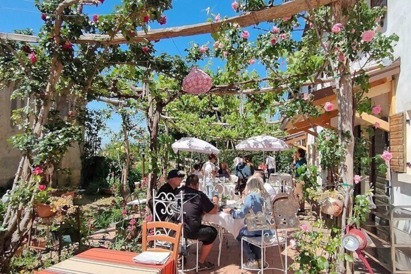An outdoor café with tables and chairs, surrounded by a garden with climbing plants and flowers. 1
