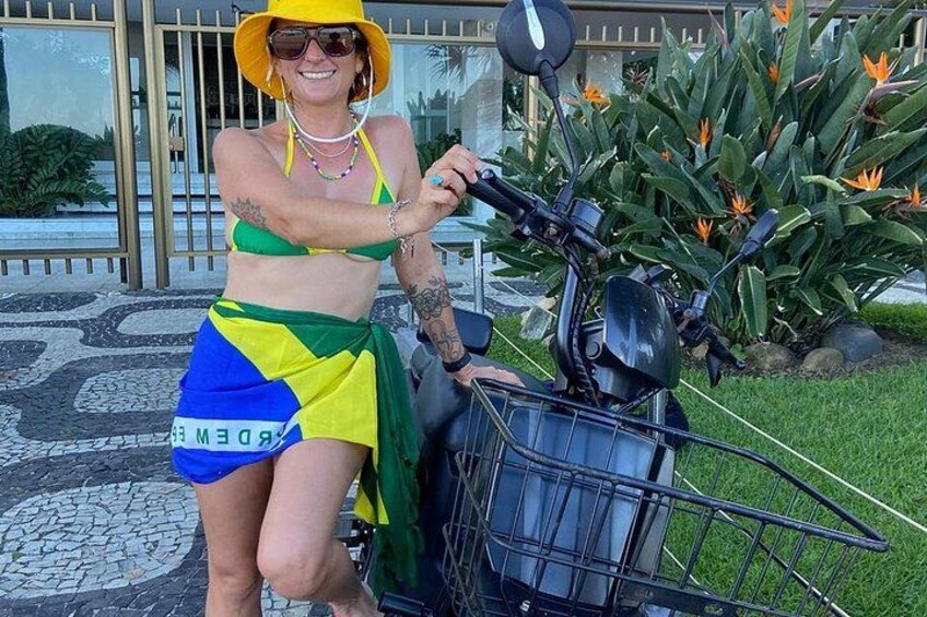 A person in a green, yellow, and blue outfit sitting on a scooter with a basket, in front of a building with a glass door and a garden with plants. 5