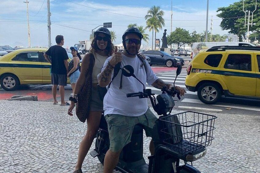 A man and a woman on a scooter with a basket, a yellow taxi, and palm trees in the background. 1