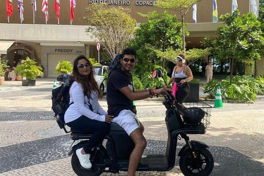 Two people on a scooter in front of a building with flags and a sign that says 'RIO DE JANEIRO COPACA'. 1