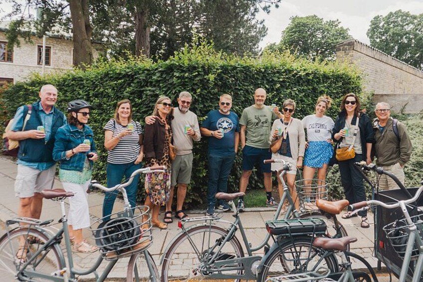 A group of people standing outdoors with bicycles, a hedge, and a building in the background. 2