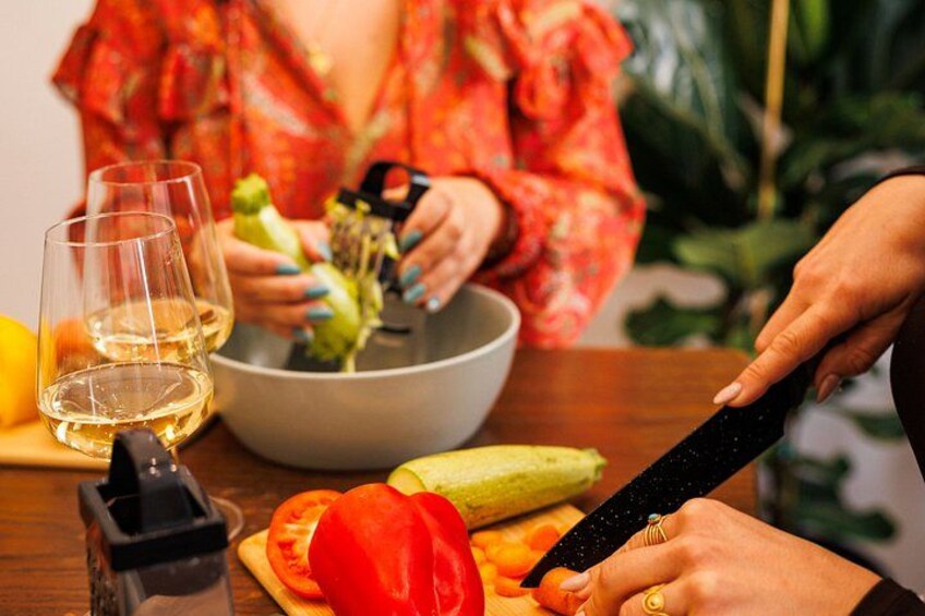 A person slicing a tomato on a cutting board with a knife, a bowl of vegetables, and two glasses of white wine on a table. 5
