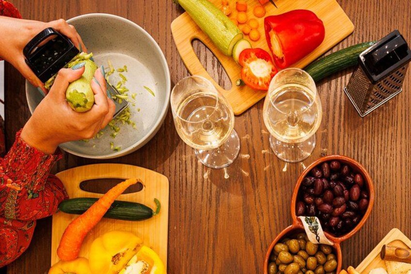 A person peeling a cucumber, a bowl of olives, a cutting board with vegetables, and two glasses of white wine. 4