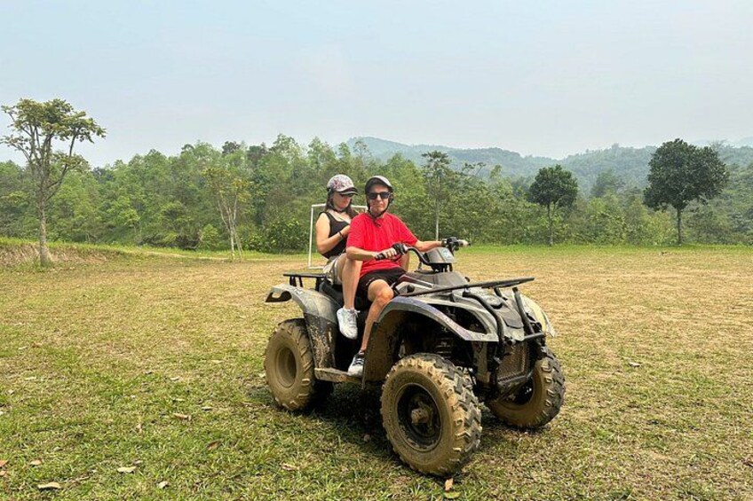 Two people on an ATV in a grassy field with trees and hills in the background. 3