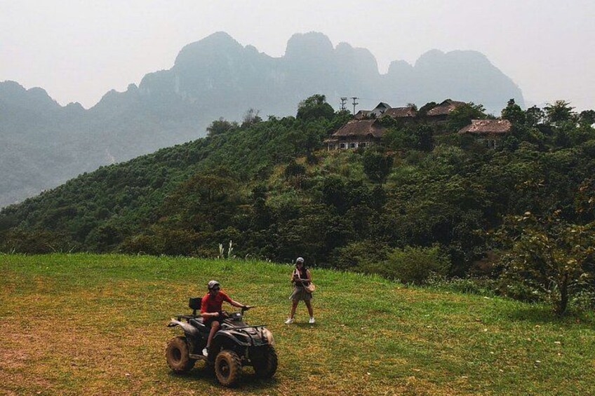 A person riding a quad bike on a grassy field with a hilly landscape and houses in the background. 5