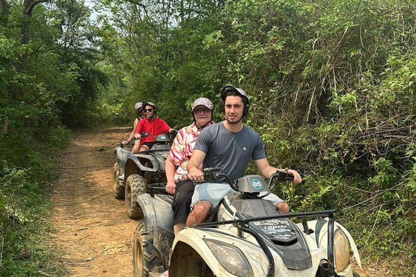 Three people on all-terrain vehicles on a dirt path surrounded by dense greenery. 4