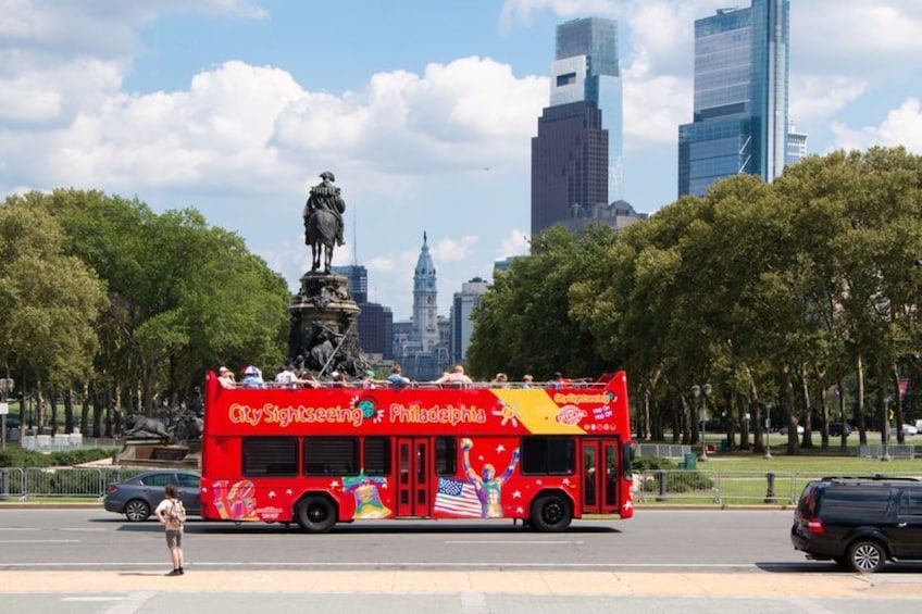 A red double-decker bus with 'City Sightseeing Philadelphia' written on it, parked in a city square with a statue and modern buildings in the background. 1