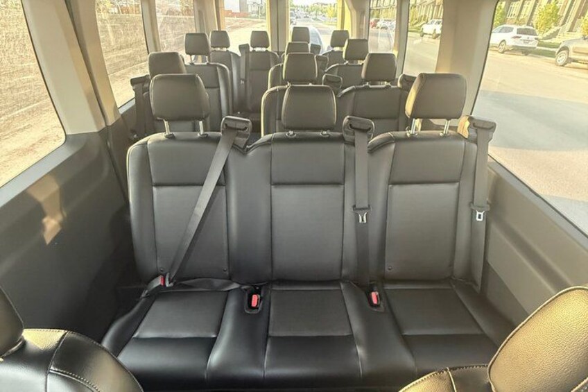 A bus interior with multiple rows of black leather seats, seat belts, and a window view of a street. 3